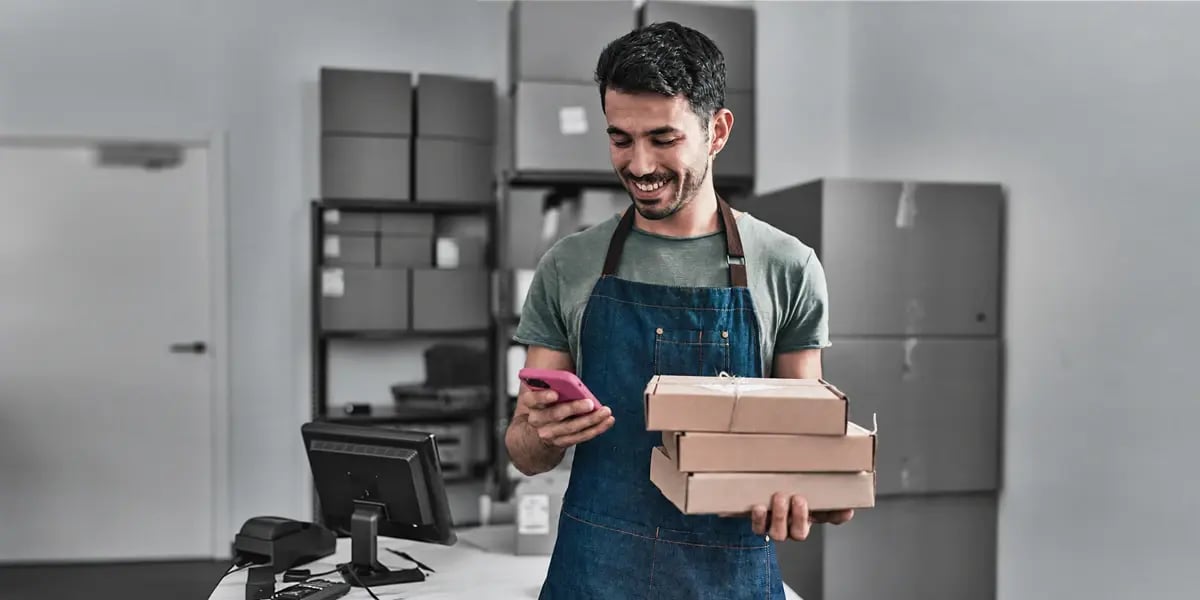 A retailer checks an order on his phone while holding a stack of parcels