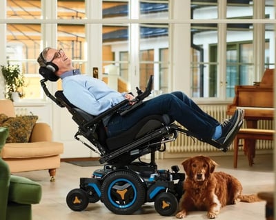 Man relaxing in a reclined Permobil powered wheelchair in a bright living room, with a dog lying beside him