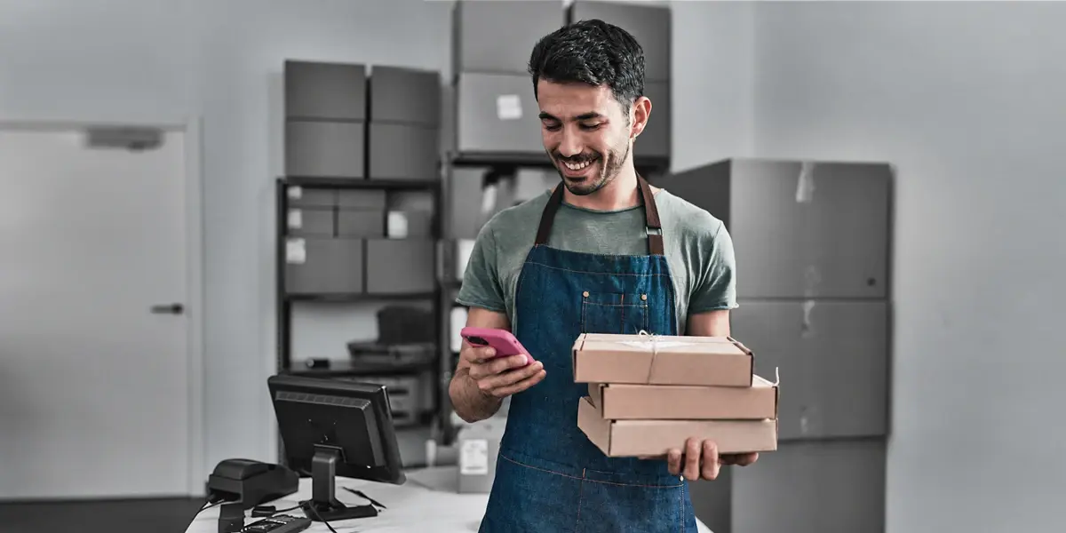 A retailer checks an order on his phone while holding a stack of parcels
